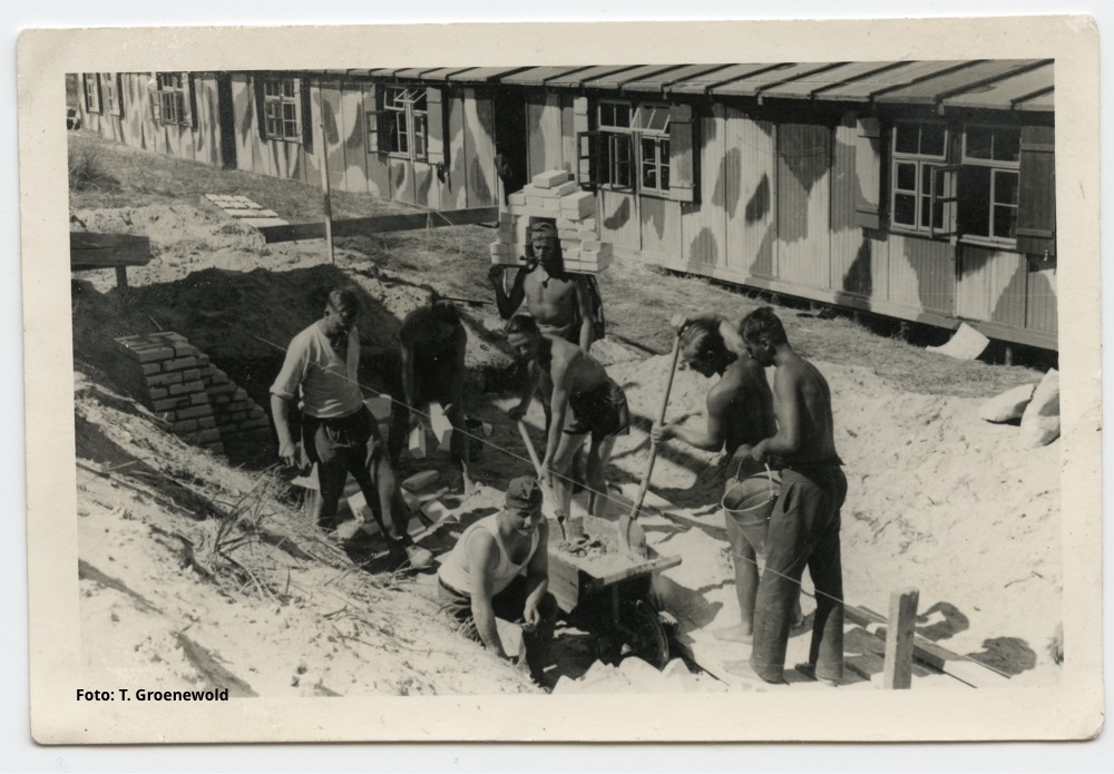 Op een Waddeneiland zijn militairen met bakstenen aan het werk voor de met camouflagepatroon geverfde houten barakken. Vanaf 1943 waren deze bijna allemaal vervangen door bunkers. - Foto: T. Groenewold