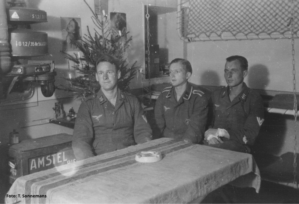 Viering van kerst in een bunker onder de grond op Schouwen. Links is boven een kist bier een gaszuiveringsinstallatie zichtbaar. Rechts aan ijzeren kettingen drie bedden waarvan de bovenste opgeklapt. - Foto: T. Sonnemans